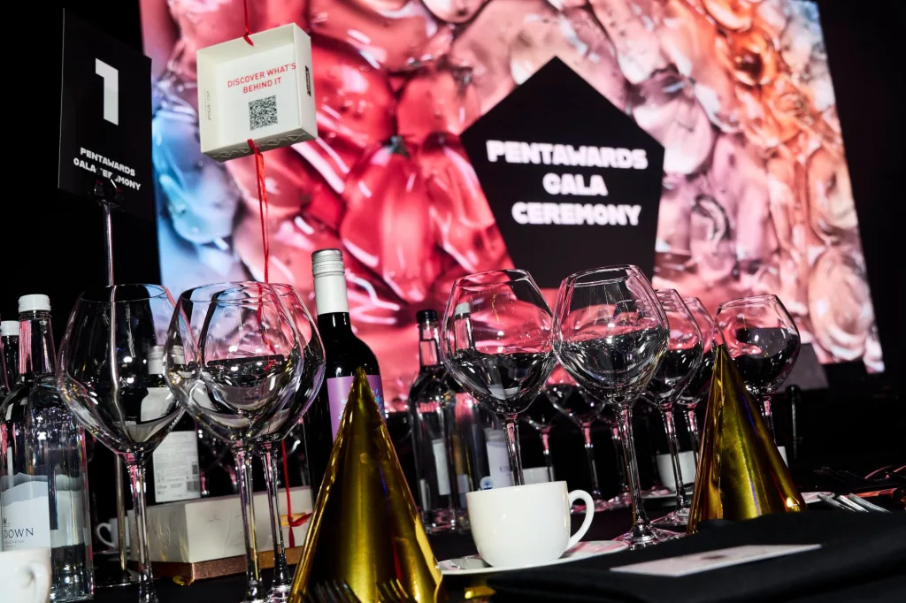 Tavolo della cerimonia di gala dei Pentawards 2026 con calici, bottiglie e cappellini dorati davanti allo schermo
